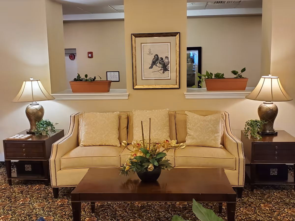 A cozy living room area with a beige sofa adorned with three patterned cushions. On either side of the sofa are dark wooden side tables, each holding a decorative lamp and a small green plant. A wooden coffee table with a floral arrangement sits in front of the sofa. The wall behind the sofa features a framed picture of three birds and two rectangular openings with potted plants on the ledges.