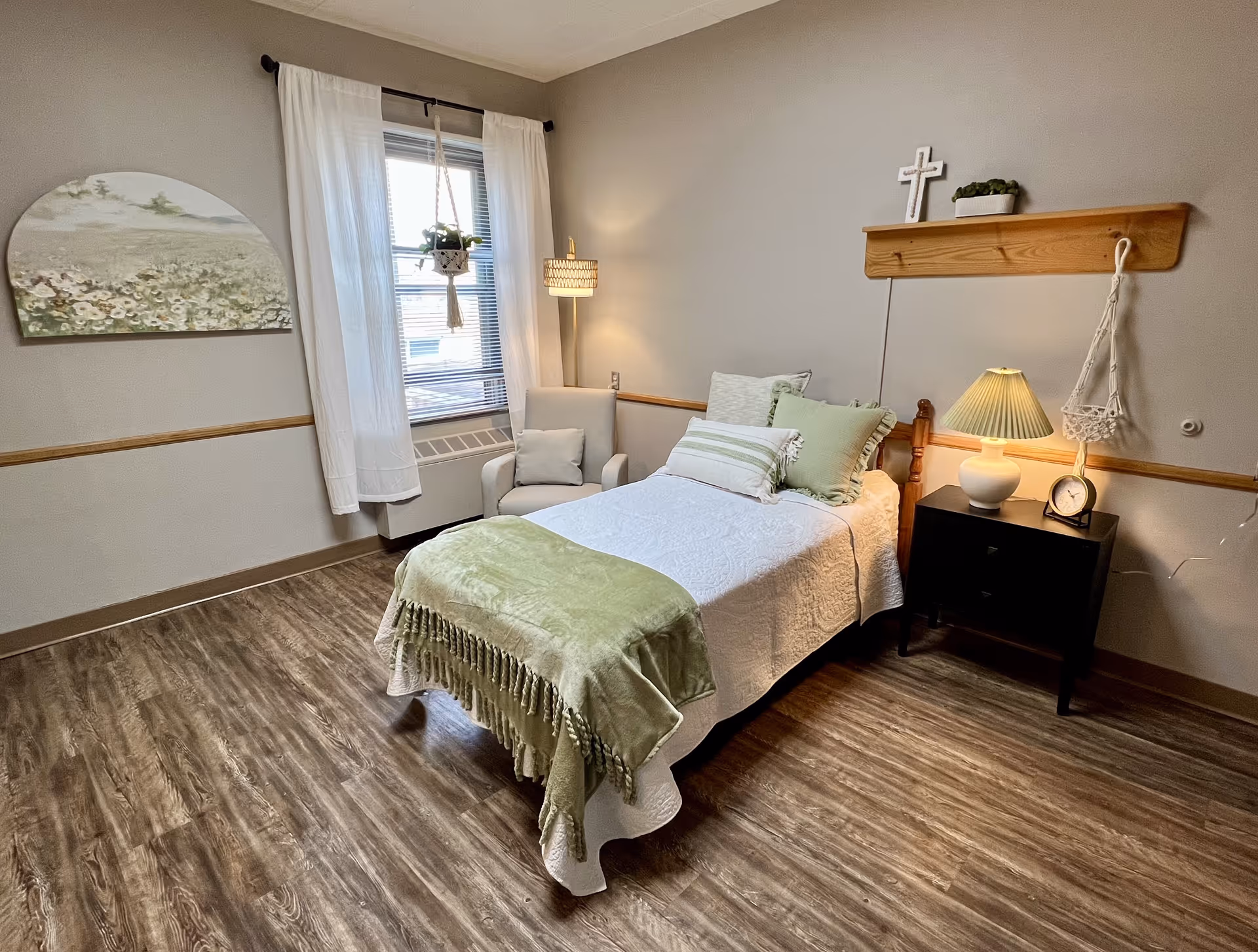 Well-lit single-occupancy bedroom with a neatly made twin bed, nightstand and lamp, armchair by a curtained window, and wall decor including a small shelf and cross.