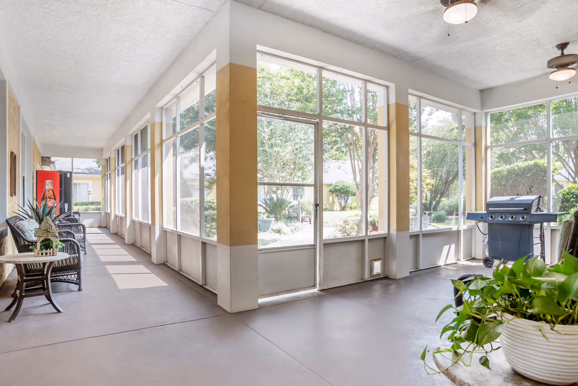 A bright, screened-in porch area with large windows overlooking a garden. The porch has wicker chairs with cushions, a small table with a potted plant, a barbecue grill, ceiling fans, and a Coca-Cola vending machine at the far end.