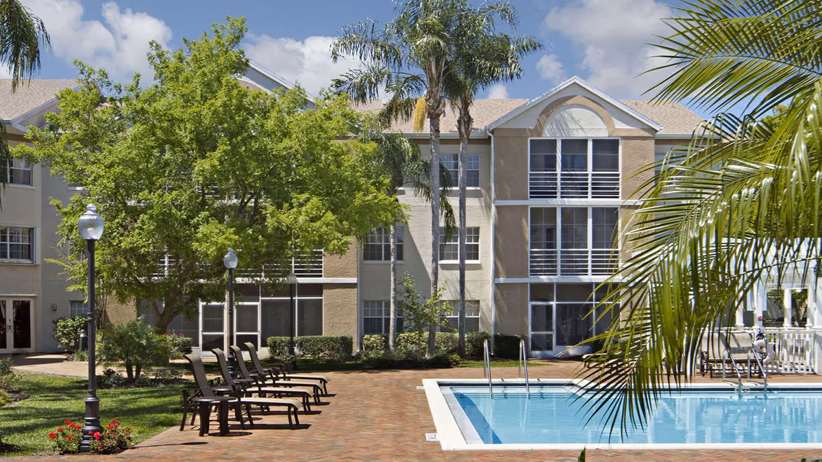 Outdoor view of a senior living facility with a swimming pool, lounge chairs, palm trees, and a multi-story building in the background under a partly cloudy sky.