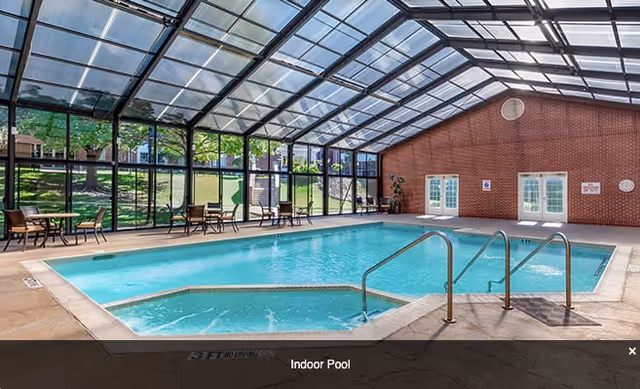 Indoor swimming pool with clear blue water inside a glass-enclosed room with a high ceiling. There are metal handrails leading into the pool, several tables and chairs along the glass walls, and a brick wall with double doors at the far end. Outside greenery is visible through the glass walls.