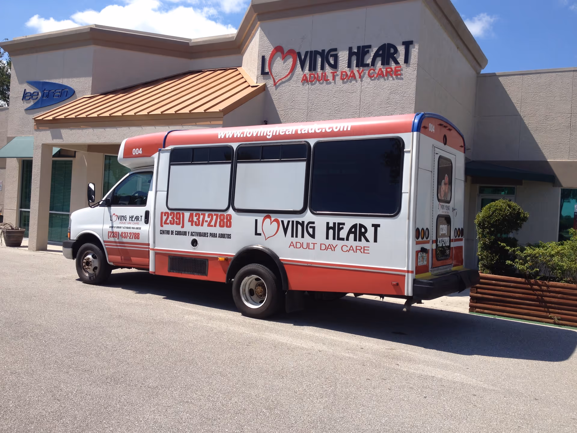 A white and red shuttle bus parked outside a building with a sign that reads Loving Heart Adult Day Care. The bus has the Loving Heart Adult Day Care logo, phone number, and website on its side. The building has a beige exterior with a small awning and some greenery nearby.