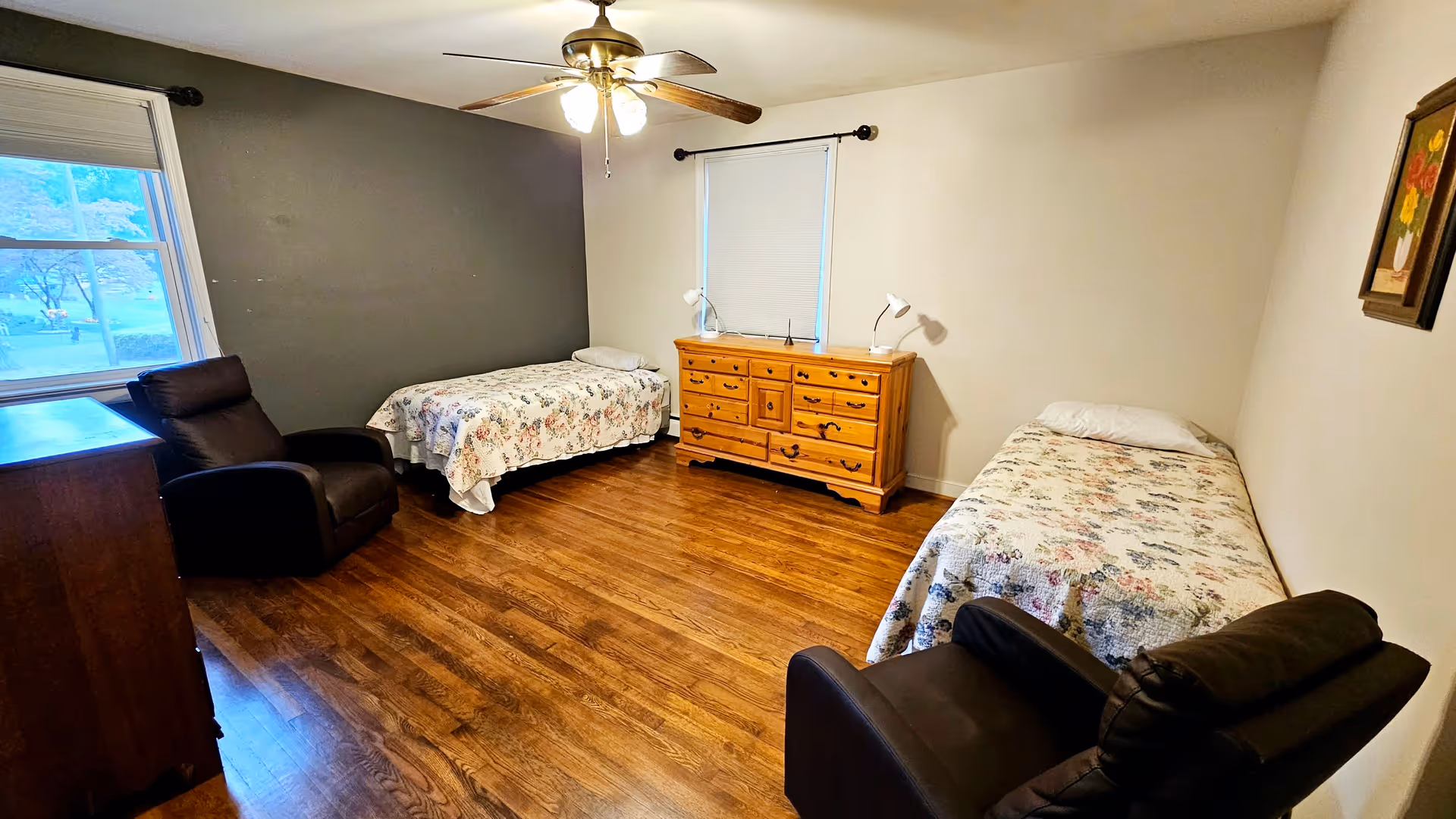 A bedroom with two single beds covered in floral bedspreads, two black armchairs, a wooden dresser with two lamps on top, a window with a closed blind, a ceiling fan with lights, and a framed painting on the wall.