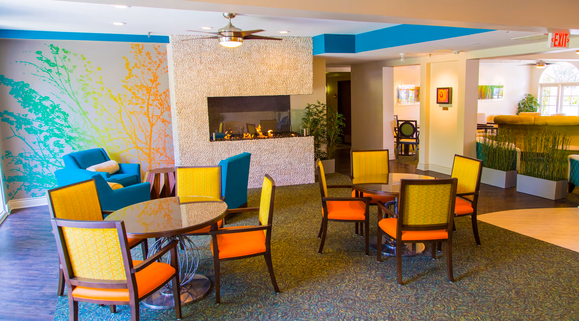 A cozy senior living common area with round tables and chairs featuring yellow and orange upholstery. There is a modern fireplace with a stone surround in the center, a ceiling fan above, and a colorful wall mural with tree branches on the left. Comfortable blue armchairs with white pillows are placed near the mural. The space is well-lit with natural light coming from large windows on the right.