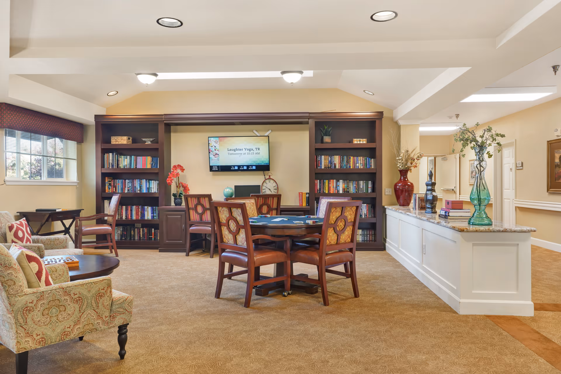 A cozy common area in a senior living facility with a round table and four chairs in the center. Behind the table are two tall bookshelves filled with books and decorative items. A flat-screen TV mounted on the wall displays a message about a laughter yoga session. To the left, there is a window with blinds and a small table with a chair. On the right side, a long counter with granite top holds several decorative vases and books. The room has beige walls, carpeted floor, and recessed ceiling lights.