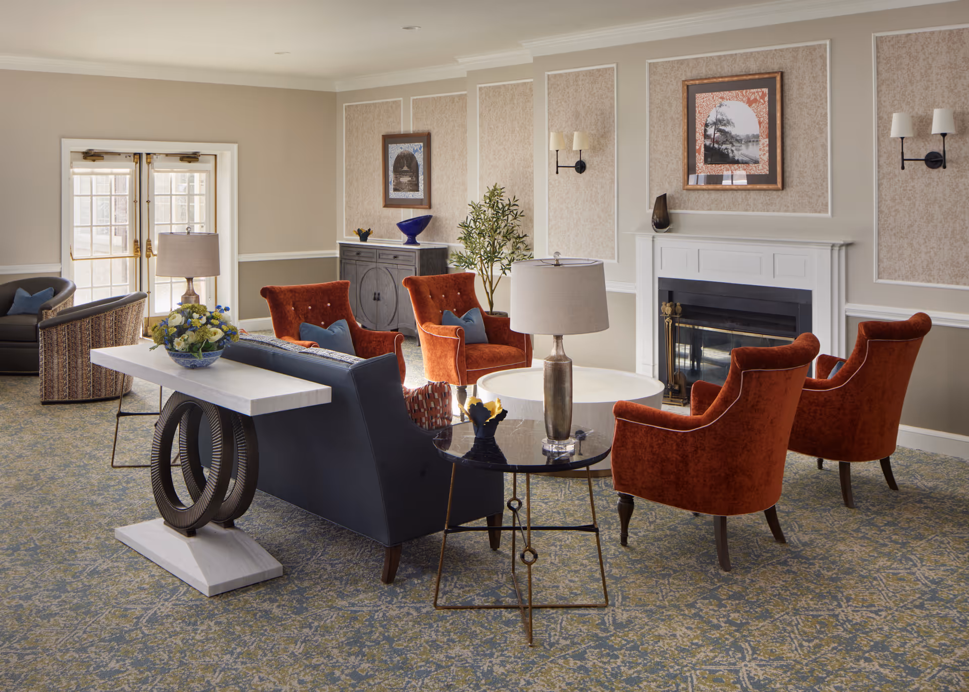 A cozy living room area in a senior living facility with a fireplace, four orange upholstered armchairs, a dark blue sofa, side tables with lamps, a console table with a flower arrangement, framed artwork on the walls, and double glass doors letting in natural light.