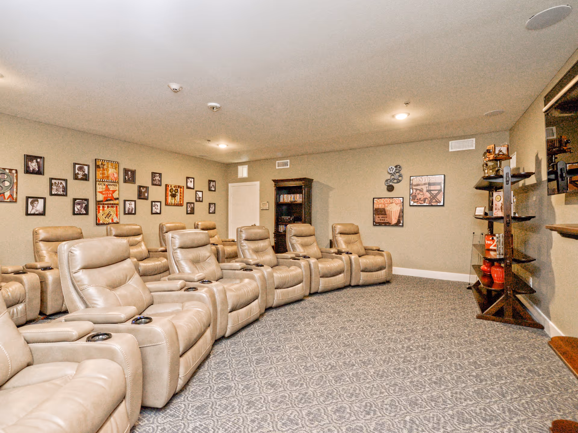 Cozy media room with multiple beige leather recliner chairs arranged in rows facing a wall-mounted screen, with framed photos and shelving on the walls.