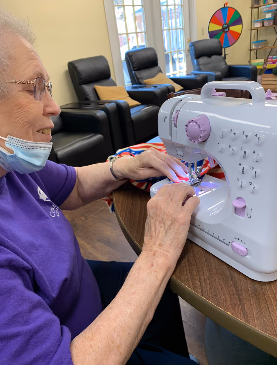An elderly woman wearing glasses and a purple Cypress Place shirt is sitting at a round table using a white and pink sewing machine. She is working on a piece of fabric with red, white, and blue colors. Behind her are black leather recliners with tan pillows, a colorful spinning wheel, and shelves with board games in a well-lit room.