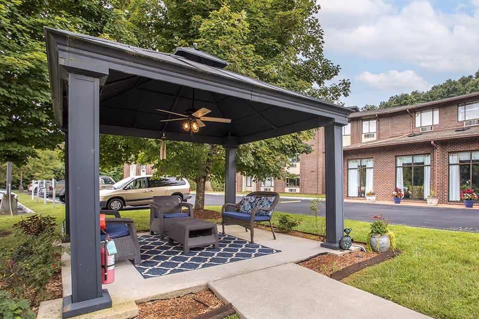 Outdoor seating area under a black gazebo with a ceiling fan and lights, featuring wicker chairs and a small table on a patterned rug, surrounded by greenery and a brick building in the background.