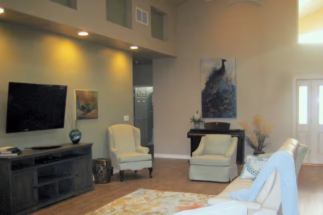 A cozy living room with a mounted flat-screen TV on the wall, a dark wooden TV stand, two cream-colored armchairs, a beige sofa with a blue throw blanket, a patterned area rug, a small table with a plant, and artwork on the walls. The room has wooden flooring and a high ceiling with recessed lighting.