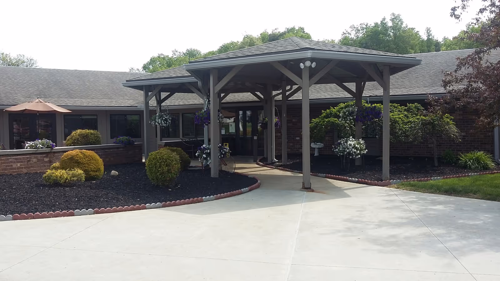 Entrance of a single-story brick building with a covered walkway supported by wooden beams. The area around the entrance is landscaped with bushes, hanging flower baskets, and a small garden bed bordered with red and white edging. There is a concrete driveway leading up to the entrance.