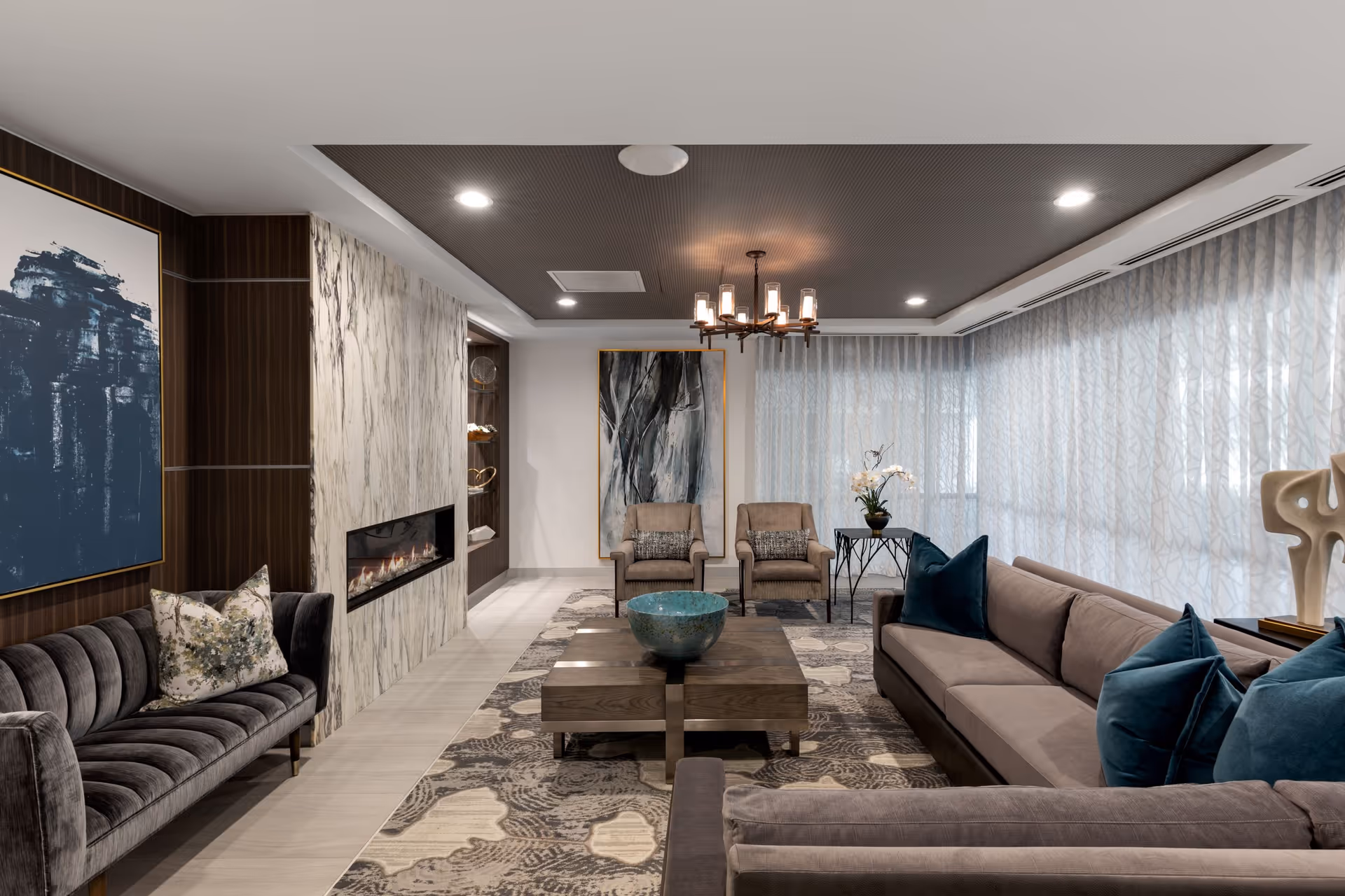 A modern living room with two gray sofas adorned with blue and patterned pillows, two beige armchairs, a wooden coffee table with a decorative bowl, a patterned area rug, a wall-mounted fireplace with a marble surround, abstract artwork on the walls, a chandelier, and large windows covered with sheer curtains.