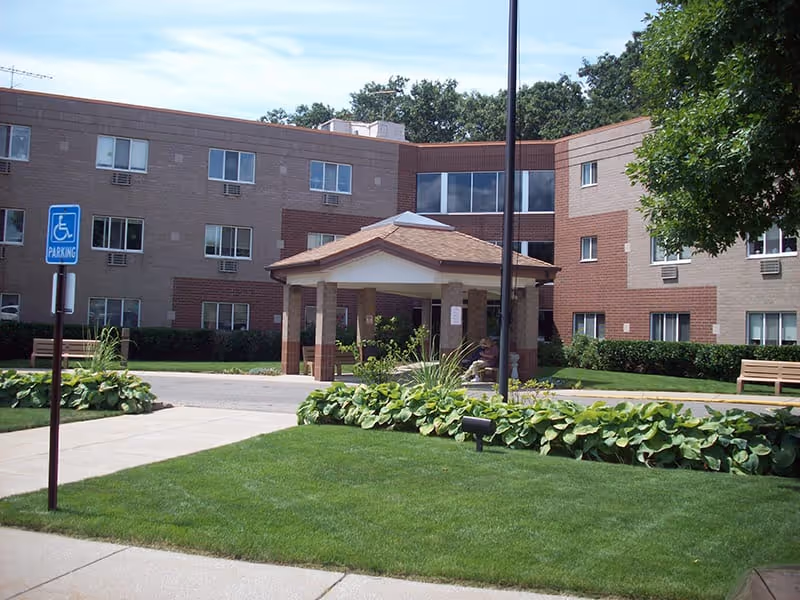 Exterior view of a multi-story assisted living facility with a covered entrance, well-maintained green lawn, bushes, benches, and a handicapped parking sign in front.
