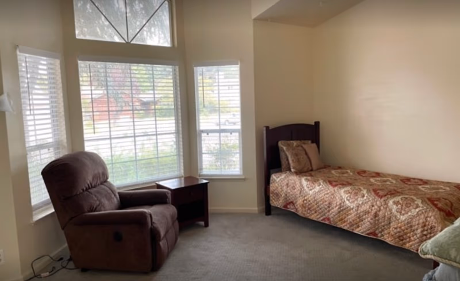 A small bedroom with a single bed covered with a patterned quilt and matching pillows, a brown recliner chair, a wooden nightstand, and large windows with white blinds letting in natural light.