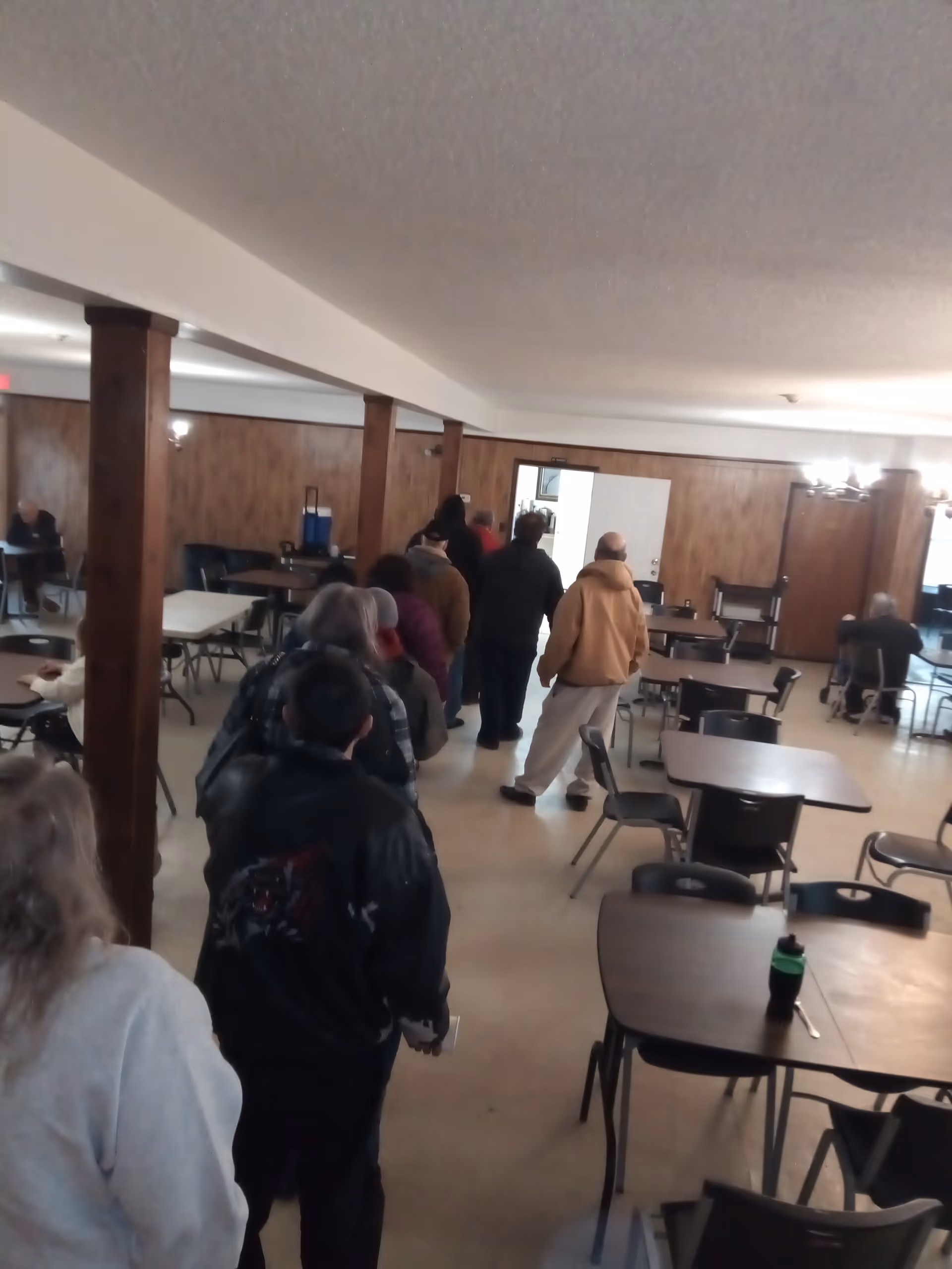 A line of people walking through a wood-paneled dining room with tables and chairs.