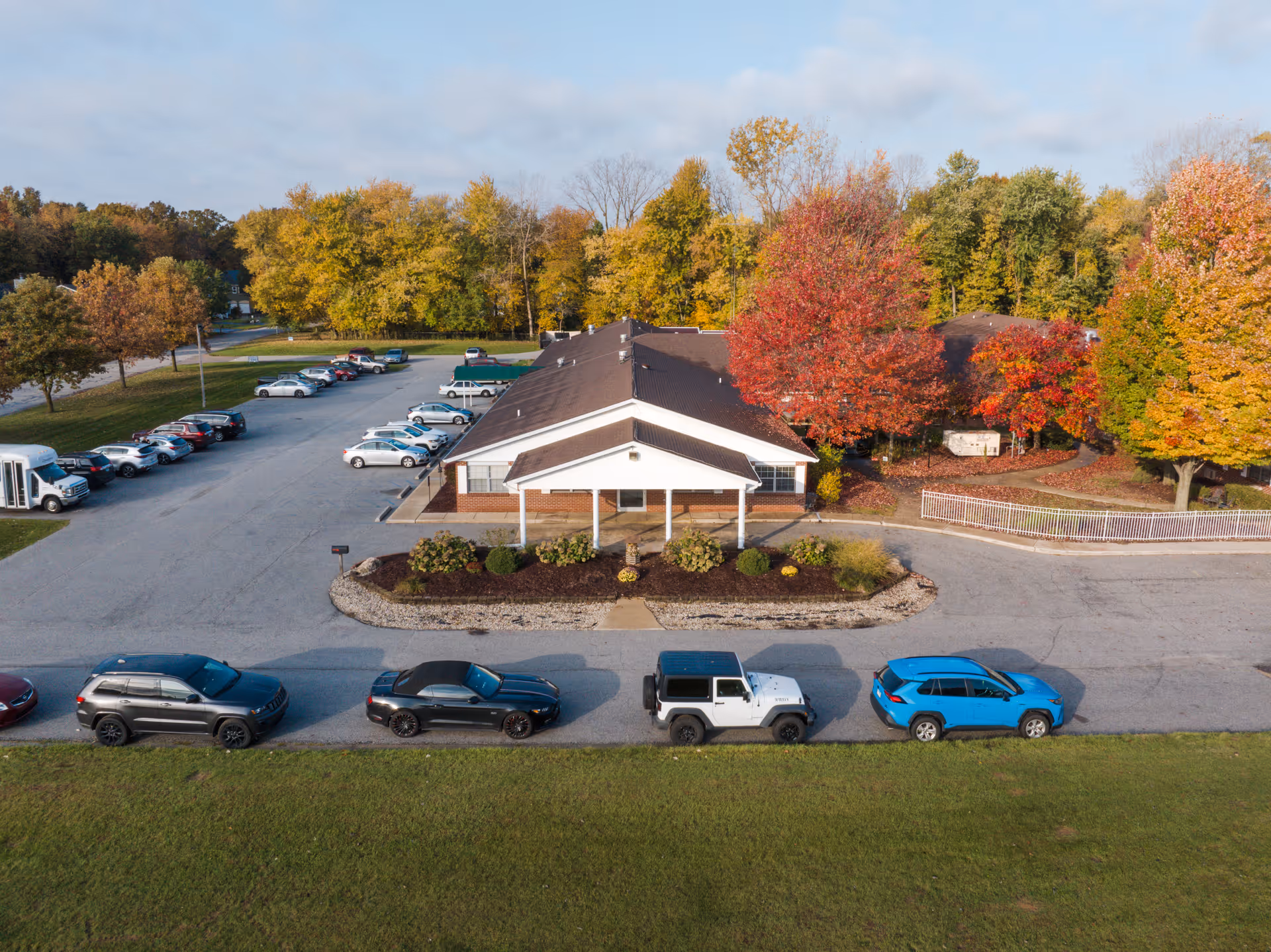 Aerial view of Chesterton Manor building with a parking lot filled with cars. The building is surrounded by trees with autumn foliage in shades of green, yellow, and red. The sky is partly cloudy.