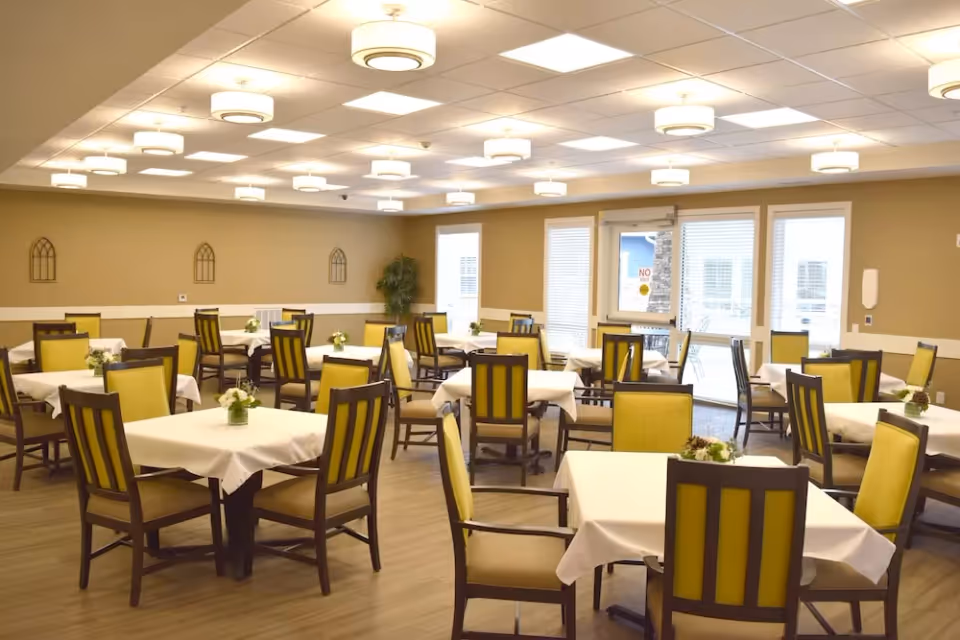 A spacious dining room with multiple square tables covered with white tablecloths, each surrounded by wooden chairs with yellow cushioned backs and seats. Small floral centerpieces are placed on each table. The room has beige walls, a light wood floor, and several ceiling lights. There are windows and a glass door at the far end of the room allowing natural light to enter.
