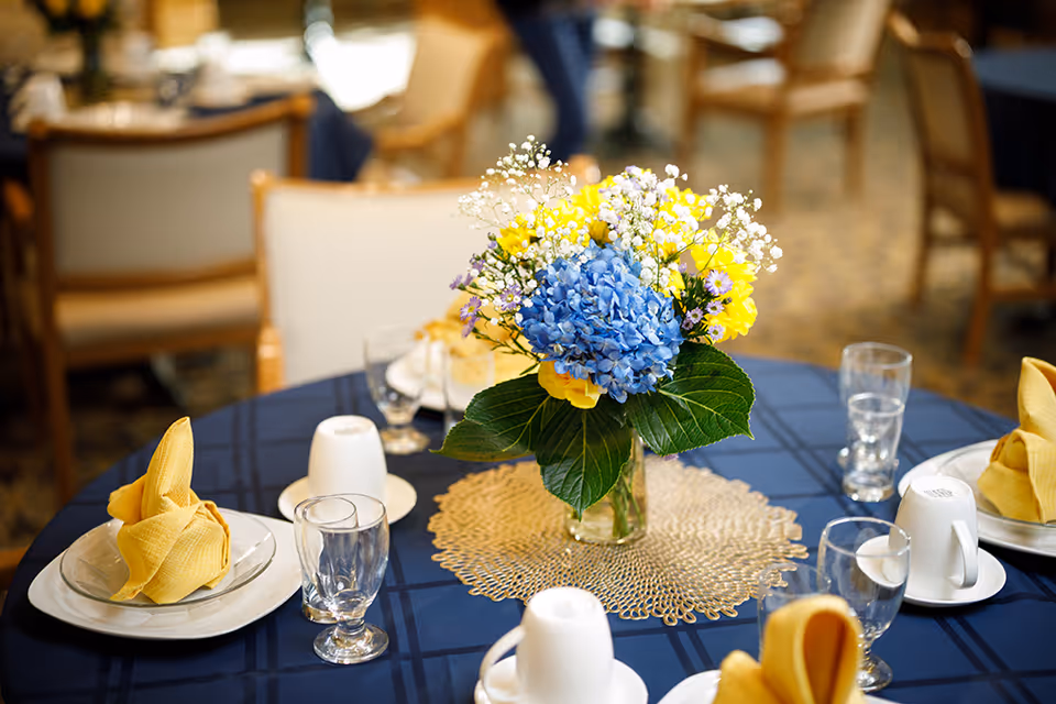 A set dining table with a blue and yellow floral centerpiece, folded yellow napkins, glasses, cups, and chairs in the background.