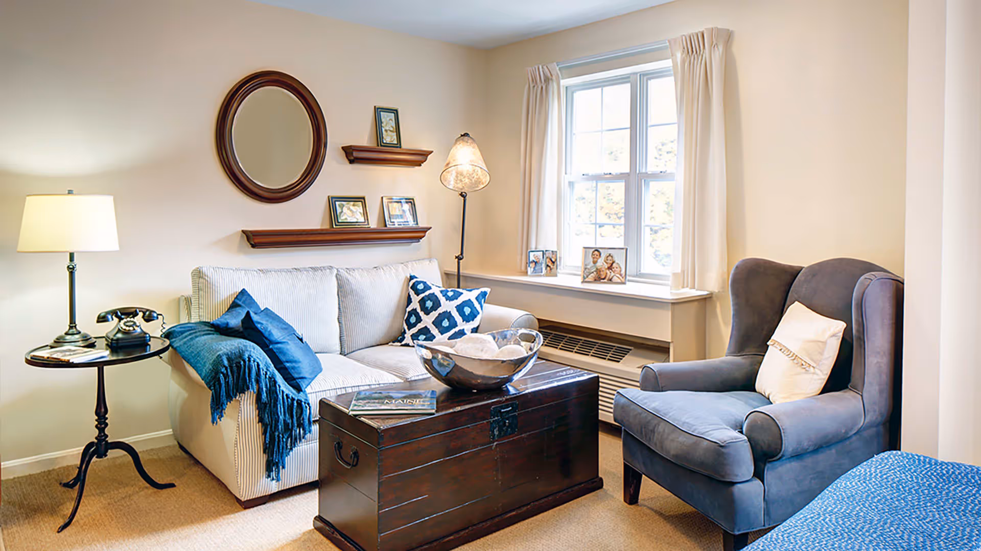 A cozy living room with a beige sofa adorned with blue and white patterned pillows and a blue throw blanket. A dark wooden chest serves as a coffee table with a decorative bowl on top. To the right, there is a comfortable blue armchair with a white pillow. A window with cream-colored curtains lets in natural light. On the left side, a small round side table holds a vintage telephone and a lamp. The wall features a round mirror and two wooden shelves with framed pictures and small decor items.