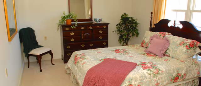 A bright bedroom with a floral bedspread and throw, wooden headboard, dresser with mirror, potted plants, and a chair.