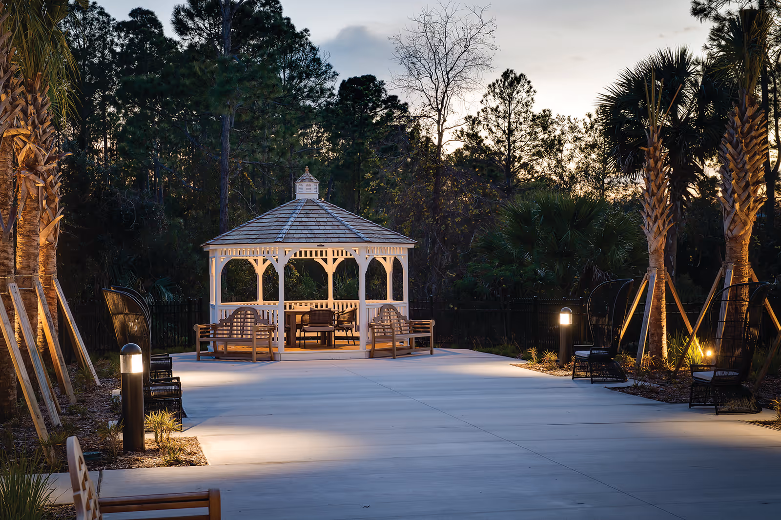 Outdoor pathway leading to a white gazebo with benches and chairs, surrounded by palm trees and other greenery, illuminated by pathway lights during dusk.