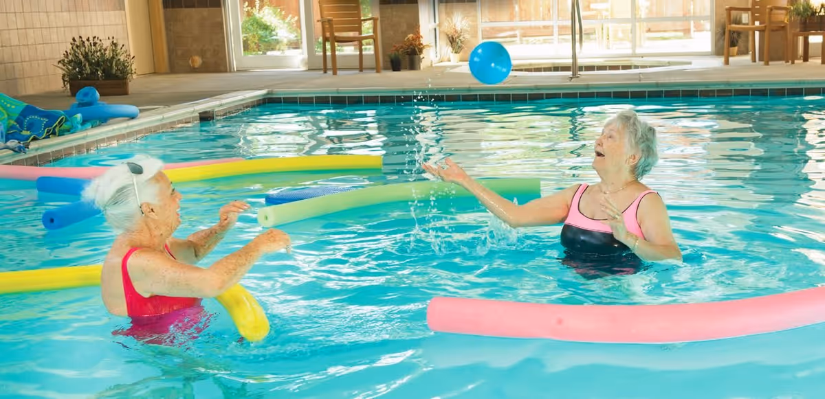 Two elderly women in swimsuits playing with a blue ball in an indoor swimming pool surrounded by colorful pool noodles. The pool area has large windows letting in natural light and a chair near the poolside.