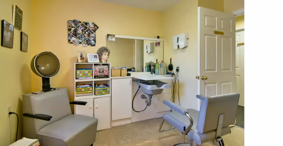 A small hair salon room with two gray salon chairs, a hair dryer, a sink with a mirror above it, and shelves holding hair care products and a mannequin head with a wig. The walls are painted light yellow, and there are photos and decorations on the walls.