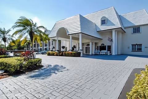Exterior view of a senior living facility with a large covered entrance, white building with multiple windows, palm trees, and landscaped bushes under a clear blue sky.