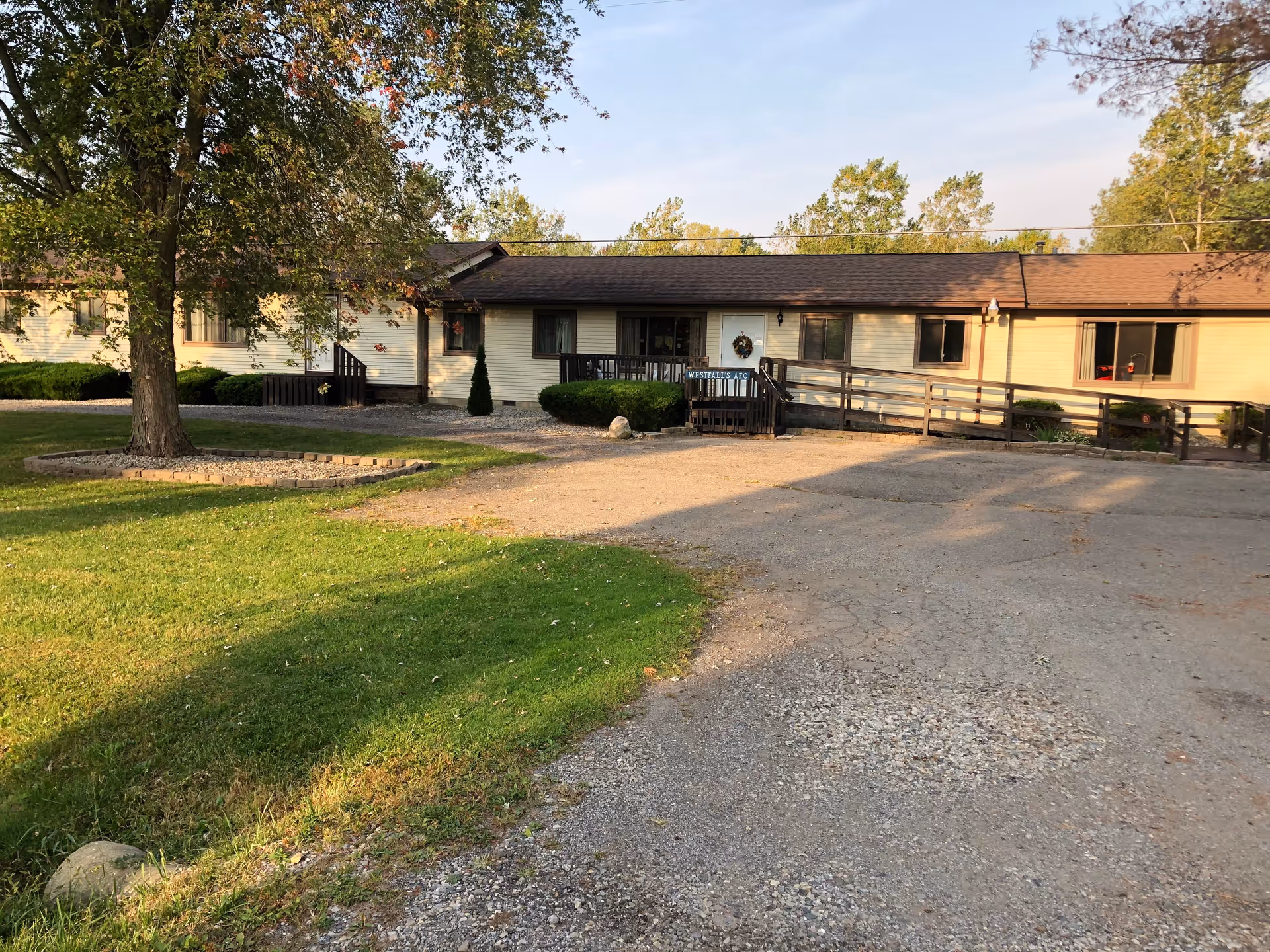 Exterior view of a single-story building with beige siding and a brown roof, surrounded by green grass and trees. There is a gravel driveway leading up to the entrance, which has a small porch with a ramp and stairs. A sign near the entrance reads 'Westfall AFC'.