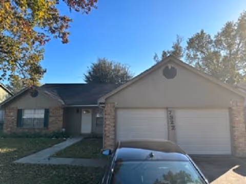 Single-story residential house with a two-car garage, brick and siding exterior, a driveway with a parked car, and trees with autumn foliage under a clear blue sky.