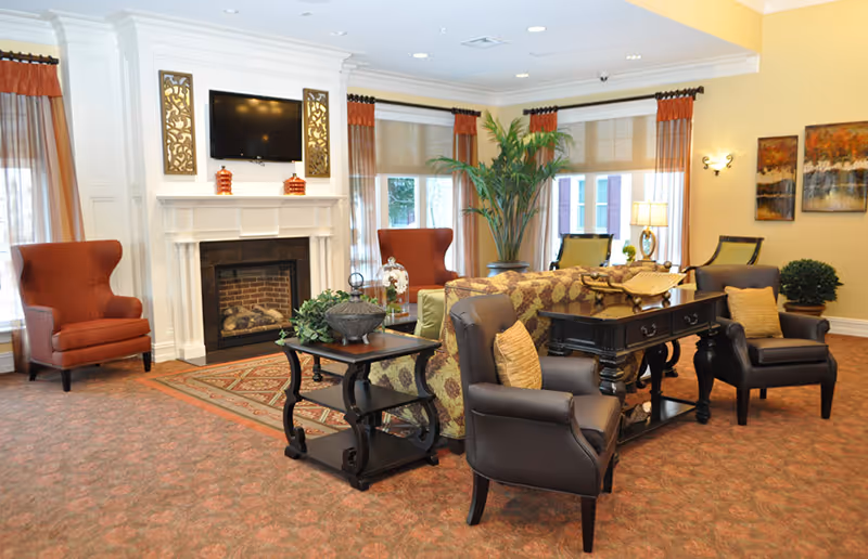 A cozy living room area in a retirement community featuring a white fireplace with a mounted flat-screen TV above it, two red armchairs on either side, a patterned sofa, two dark leather armchairs with beige cushions, a wooden side table with decorative plants, large windows with sheer and orange curtains, and warm lighting with wall sconces and a table lamp.