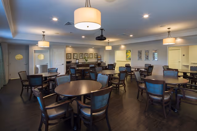 Well-lit senior dining room with multiple round wooden tables and chairs, pendant lights, and a service counter at the back.