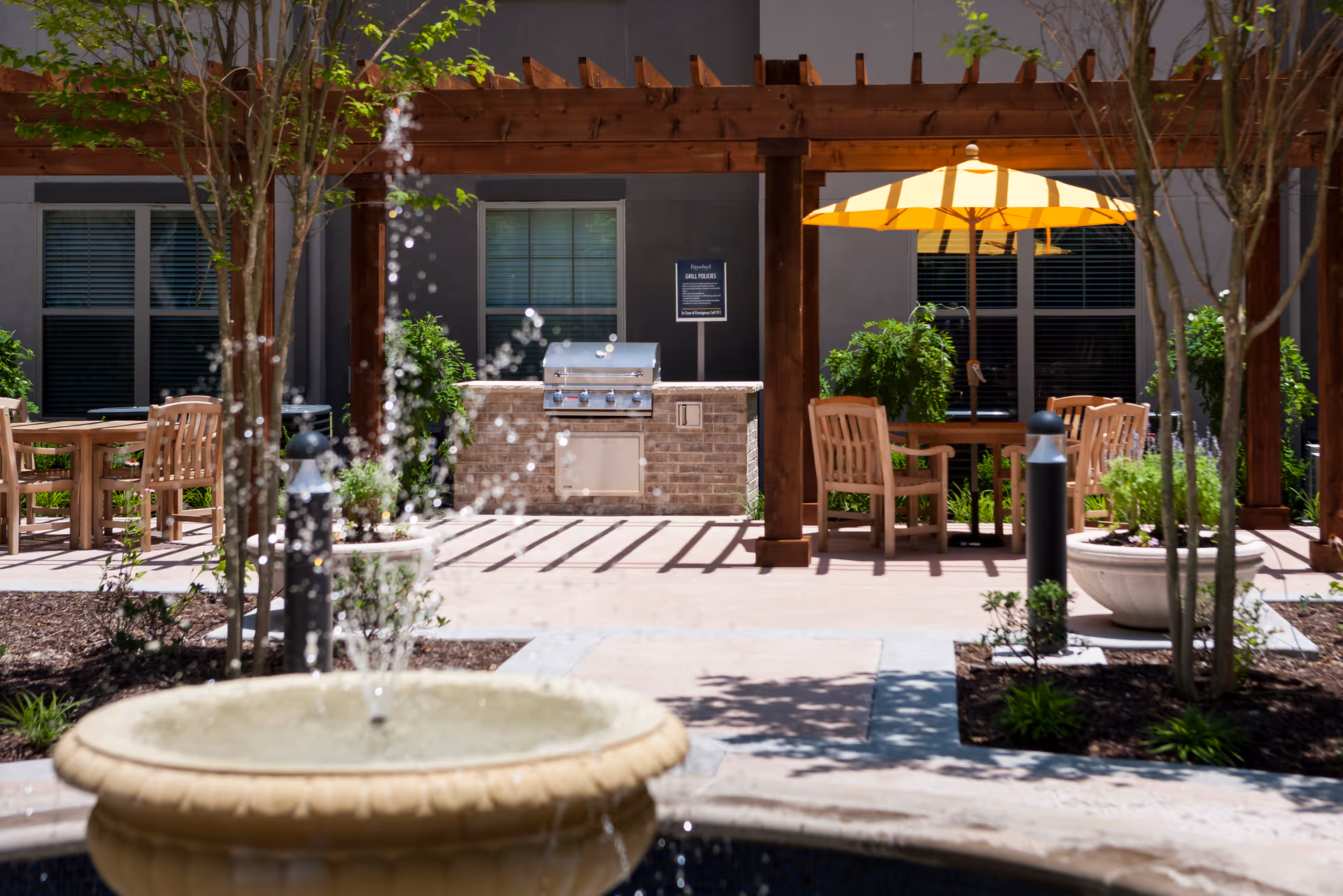 An outdoor patio with wooden tables and chairs under a pergola and umbrella, near a grill.