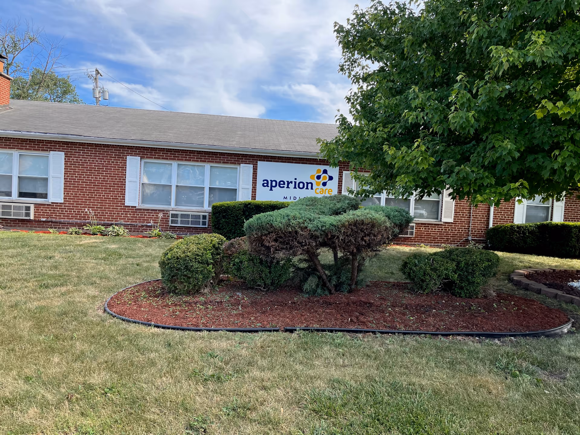 Brick single-story senior living building with an 'aperion care' sign, lawn, trimmed shrubs, and a large tree in front.