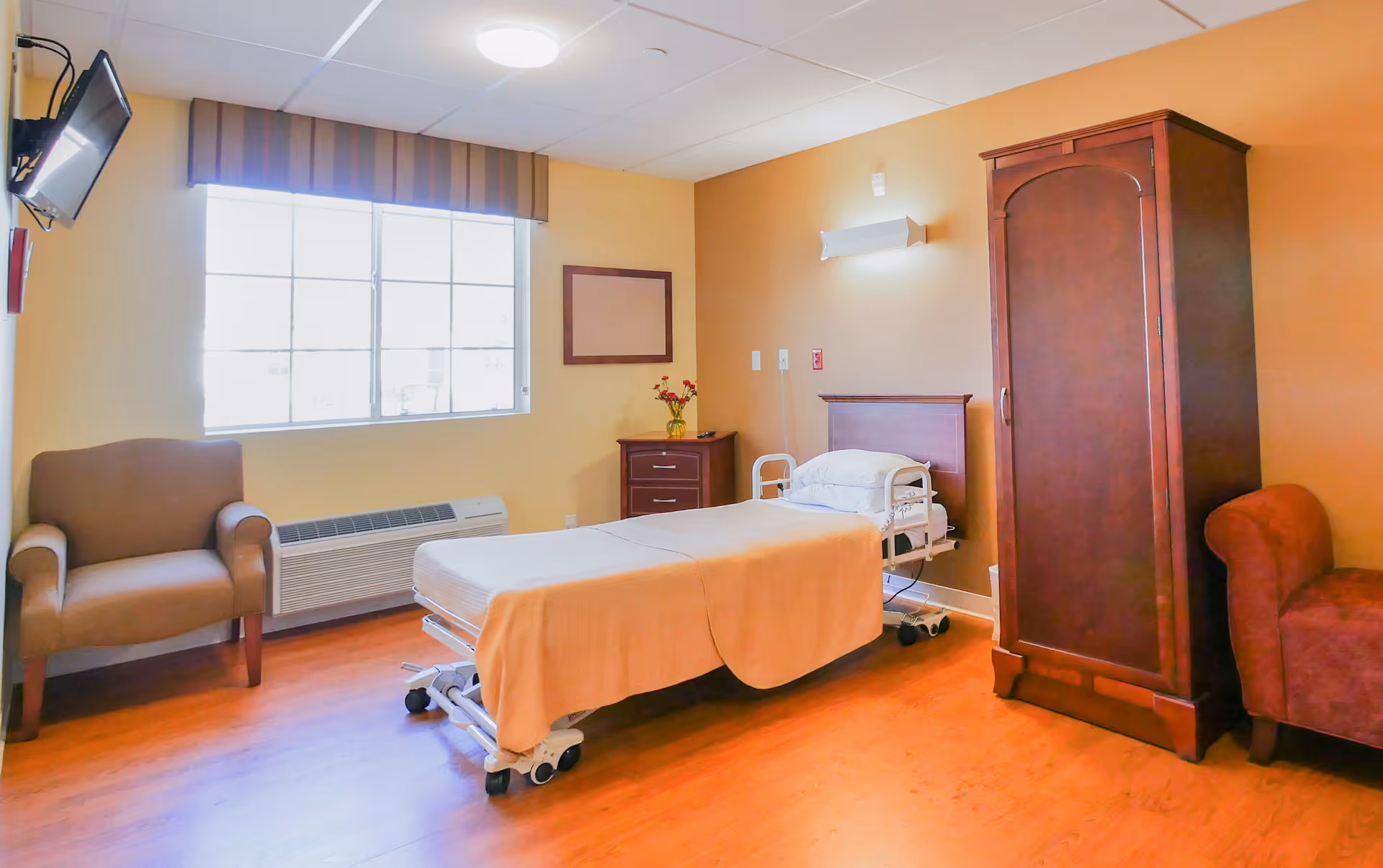A well-lit senior care room with a single hospital-style bed covered with a beige blanket, a wooden nightstand with a vase of flowers, a wooden wardrobe, a brown armchair, and a wall-mounted TV near a large window with blinds.