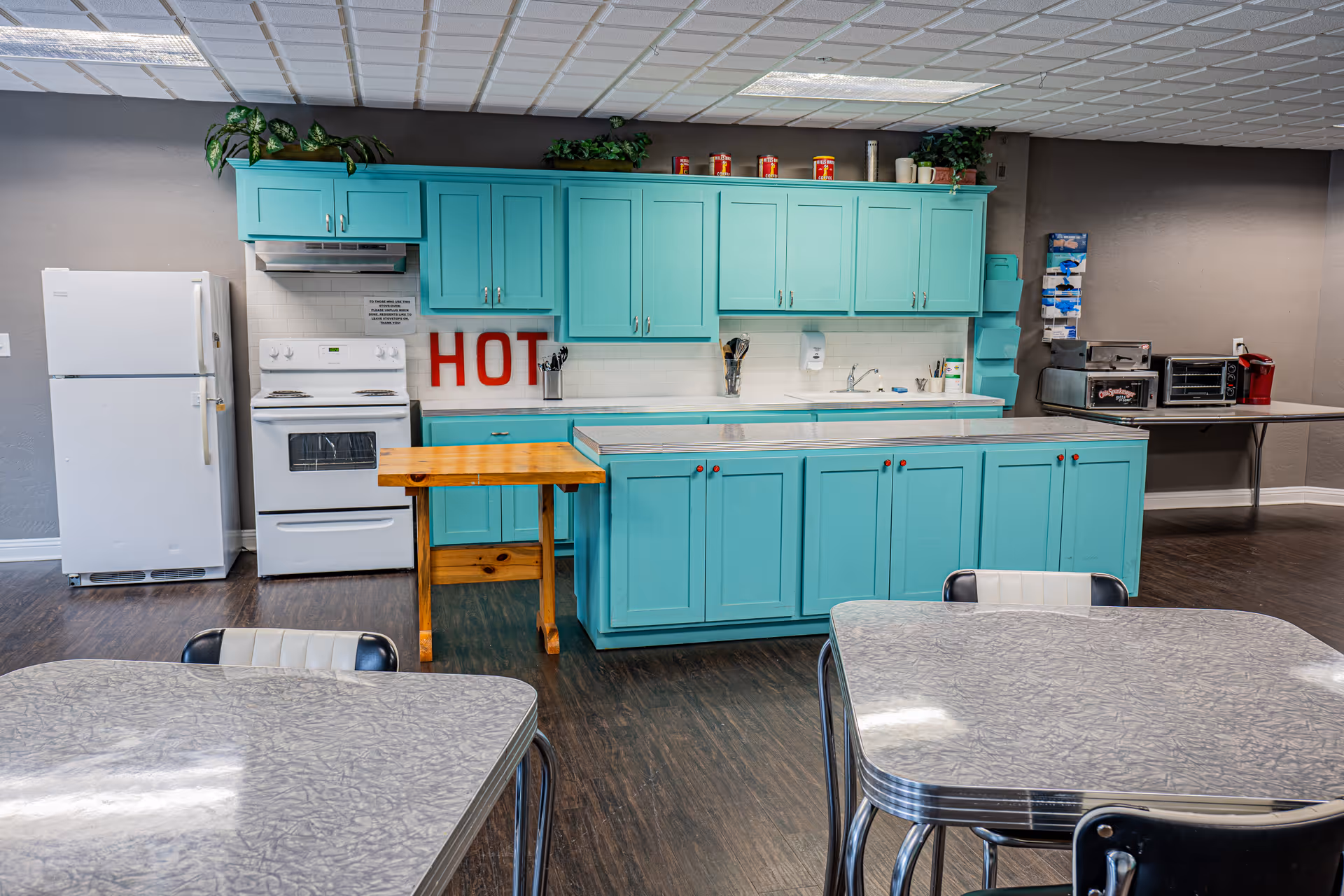 A communal kitchen area with turquoise cabinets, a white refrigerator, and a white stove. There is a small wooden table in front of the stove and two gray tables with black and white chairs in the foreground. On the back wall, there are some plants on top of the cabinets and a red sign that says 'HOT'. To the right, there is a counter with a toaster oven, microwave, and a coffee maker.