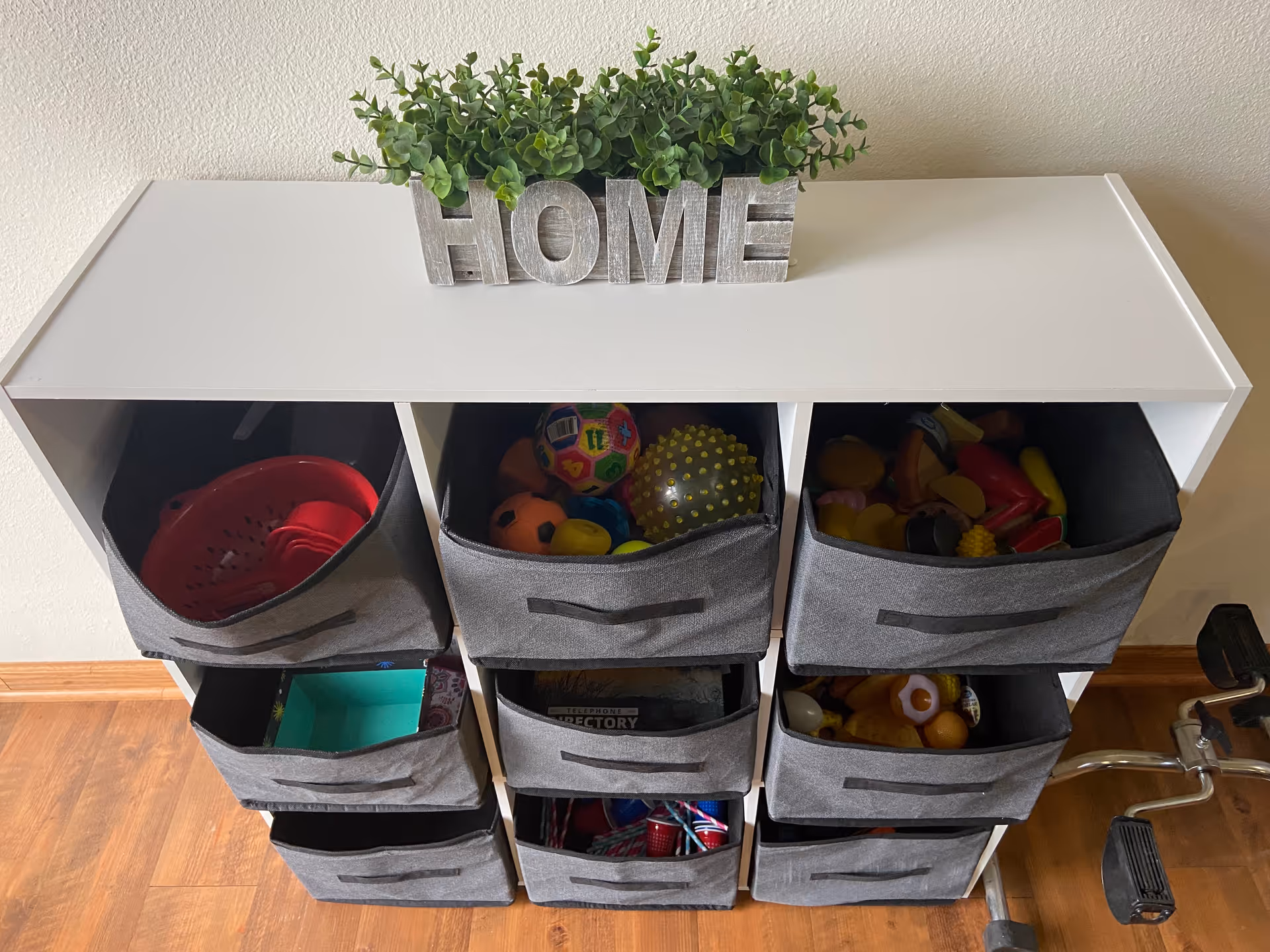 A white shelving unit with nine gray fabric storage bins filled with various toys and items. On top of the unit is a decorative plant and a wooden sign spelling 'HOME'. The unit is placed against a beige wall on a wooden floor, with part of a walker visible on the right side.