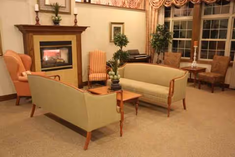 Cozy lounge in a senior facility featuring beige sofas, chairs, a fireplace, and plants.