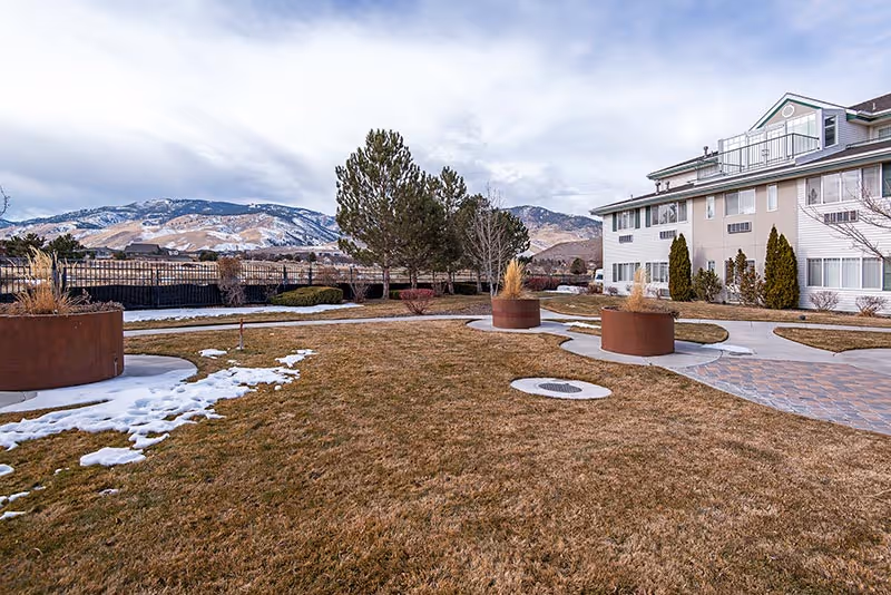 Outdoor view of a senior living facility with a large grassy area, circular planters with ornamental grasses, paved walkways, and a multi-story building on the right. Snow patches are visible on the grass, and mountains are seen in the background under a cloudy sky.