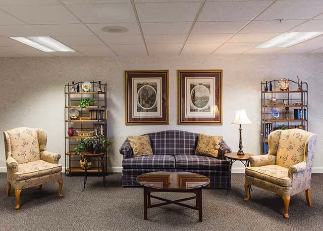 A cozy sitting area with a plaid sofa flanked by two floral armchairs, a glass-top coffee table, side tables with a lamp, framed artwork, and shelving against a wall.
