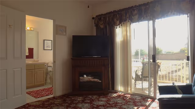 Sunlit living room with a TV on a fireplace stand, sliding glass doors to an outdoor patio, and an open doorway leading to a bathroom.