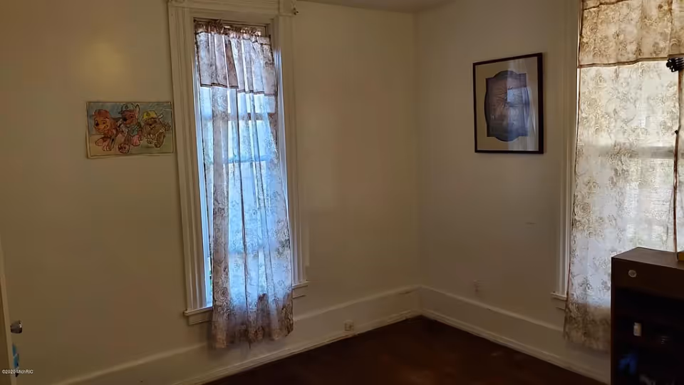 Empty room with two curtained windows, framed pictures on white walls, and a hardwood floor.