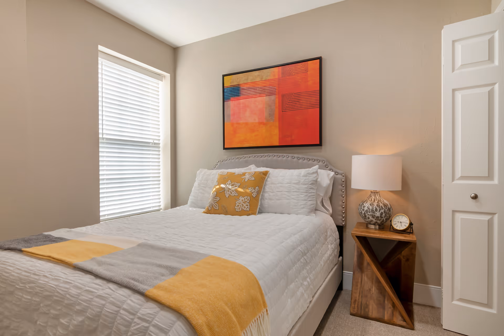 A neatly made bed with white bedding and a yellow decorative pillow in a bedroom. There is a window with white blinds on the left wall, a colorful abstract painting above the bed, and a wooden nightstand on the right side with a patterned lamp and a small clock. A white door is partially visible on the right side of the image.