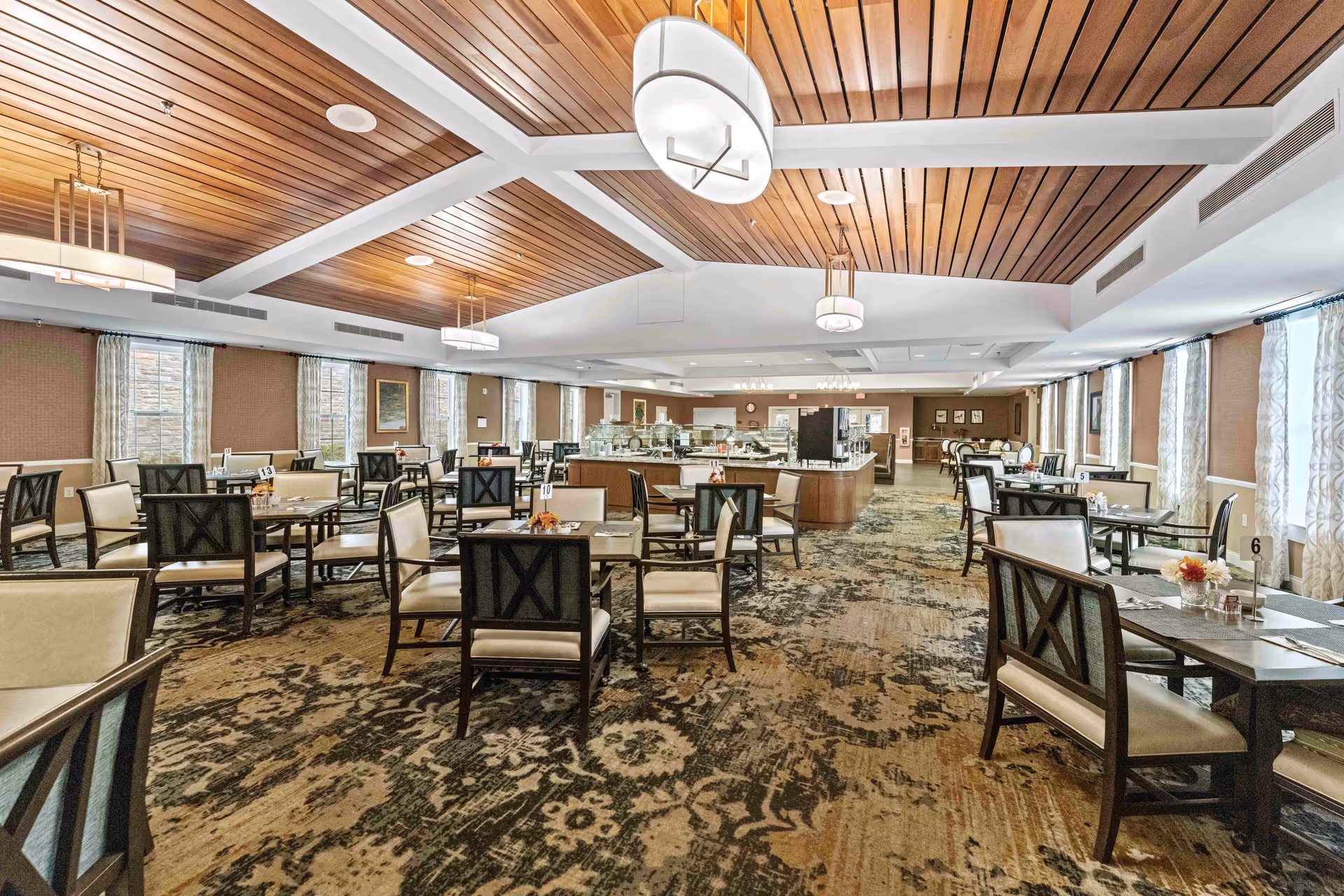 A spacious dining room with multiple tables and chairs arranged neatly. The room features a patterned carpet, large windows with curtains allowing natural light, and a wooden ceiling with modern light fixtures. In the background, there is a buffet or serving area.
