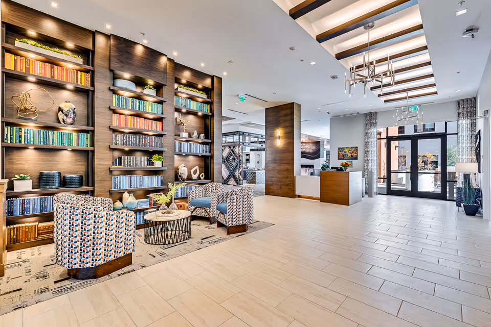 Bright and spacious assisted living facility lobby with a large wooden bookshelf filled with colorful books and decorative items on the left. Two patterned armchairs and a round coffee table are placed in front of the bookshelf on a rug. The lobby features light-colored tile flooring, modern ceiling lights with wooden beams, and a reception desk near the glass entrance doors. Large windows and glass doors allow natural light to fill the space.