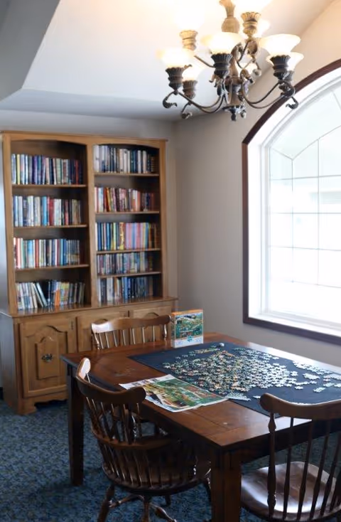 Wooden dining table with chairs and a partially completed jigsaw puzzle next to a tall bookcase and a large window under a chandelier.