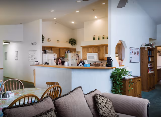 Open communal interior showing a kitchen counter with two people behind it, a dining table, and a sofa.