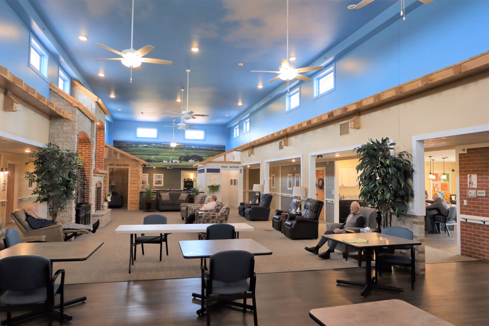 A spacious and well-lit common area in a senior living facility with a high blue ceiling, ceiling fans, and multiple windows near the ceiling. The room features comfortable seating including recliners and armchairs arranged around tables. There is a large stone fireplace on the left side, potted plants, and a mural of a countryside landscape on the far wall. Several elderly residents are seated and relaxing in the room. The area has a warm and inviting atmosphere with a mix of carpet and wood flooring.