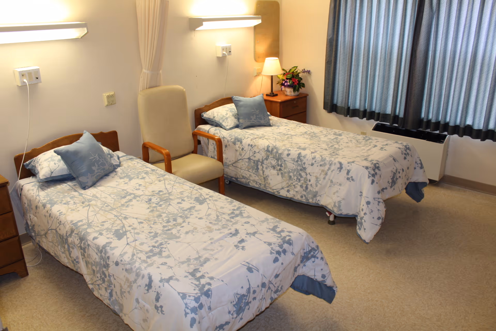 A room with two single beds covered in white and blue floral bedspreads, each with a matching pillow. Between the beds is a beige armchair with wooden armrests. There are two wooden nightstands, one with a lamp and a flower arrangement. The room has beige walls, a window with blue curtains, and wall-mounted lights above each bed.