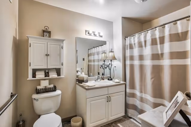 A clean and well-lit bathroom featuring a white toilet with a cabinet above it holding towels and a clock, a vanity with a sink and countertop accessories, a large mirror, a decorative lamp, and a shower with a beige and white striped curtain.